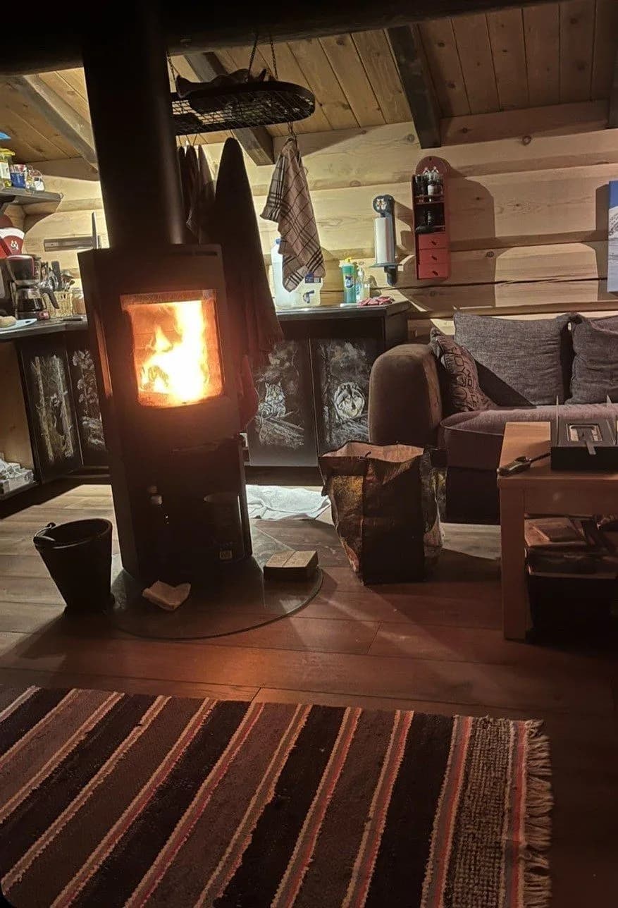 Koselig peisestue inne i anneksset med vedovn som brenner, tømmervegger og fillerye på gulvet.