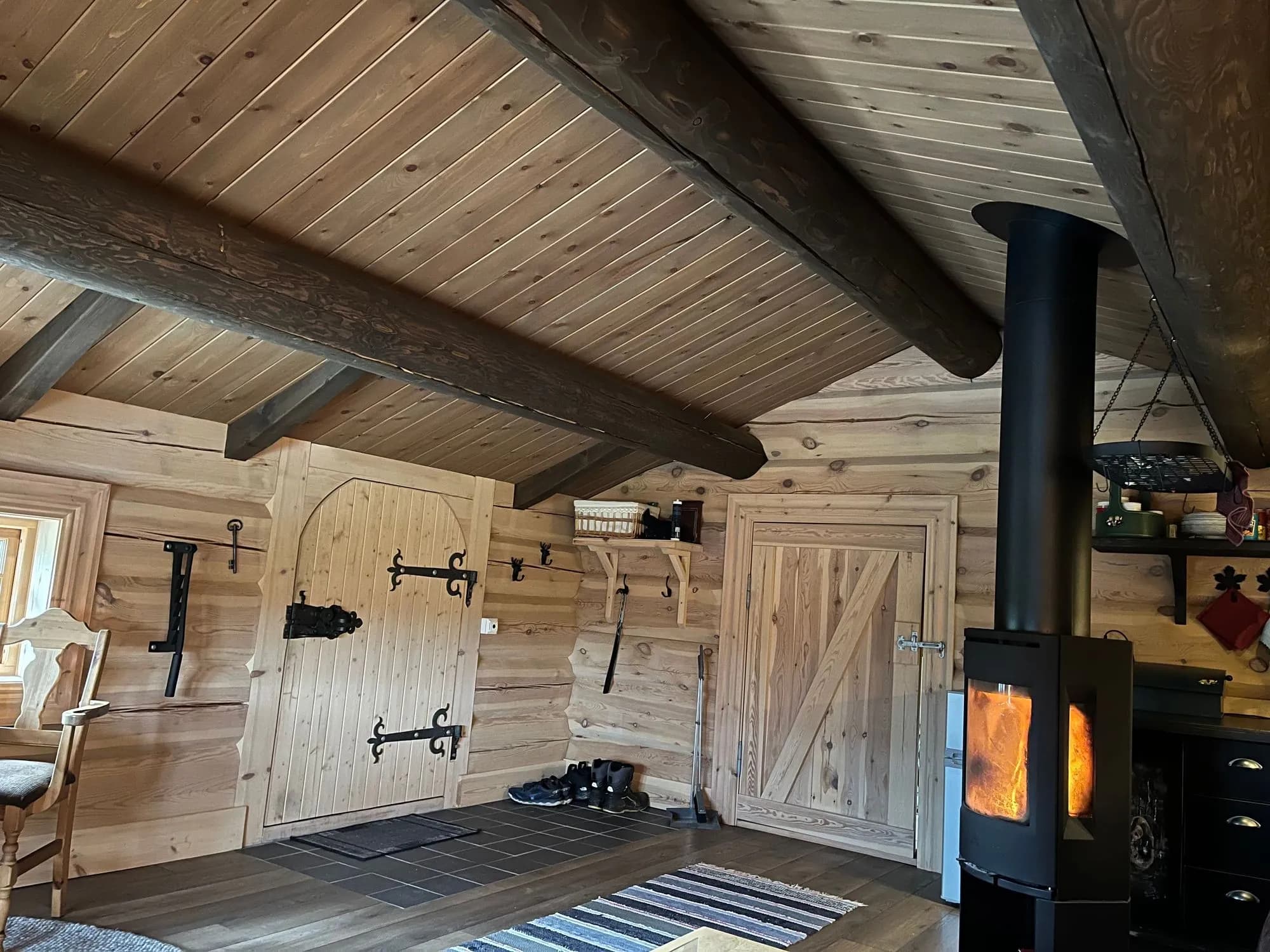 Entré i anneksset med tømmervegger, panelt skråtak og en brennende vedovn, med to tredører og fillerye på tregulvet.