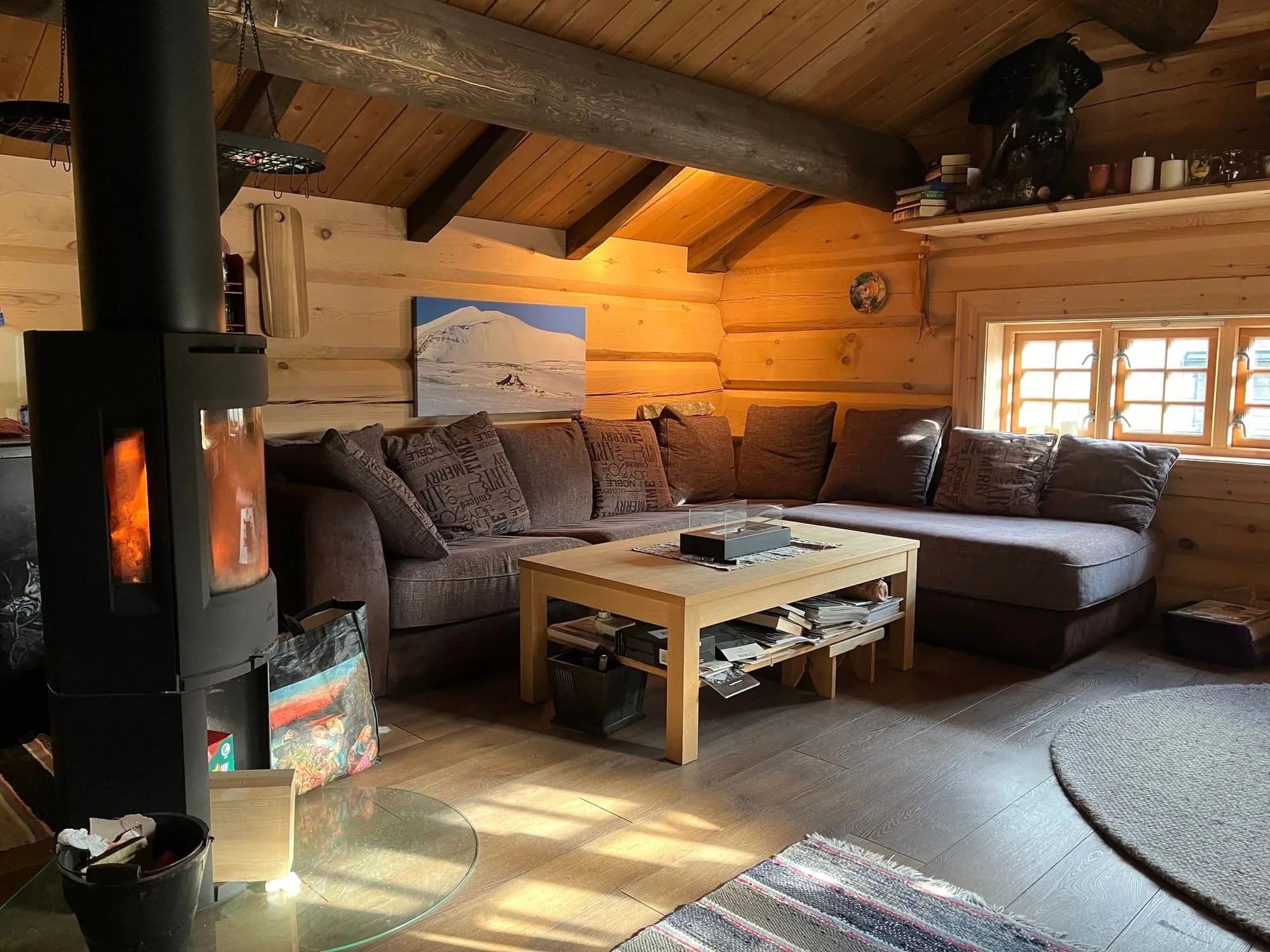 Peisestue i anneksset med brun hjørnesofa, brennende vedovn, tømmervegger og et fjellmaleri over sofaen.