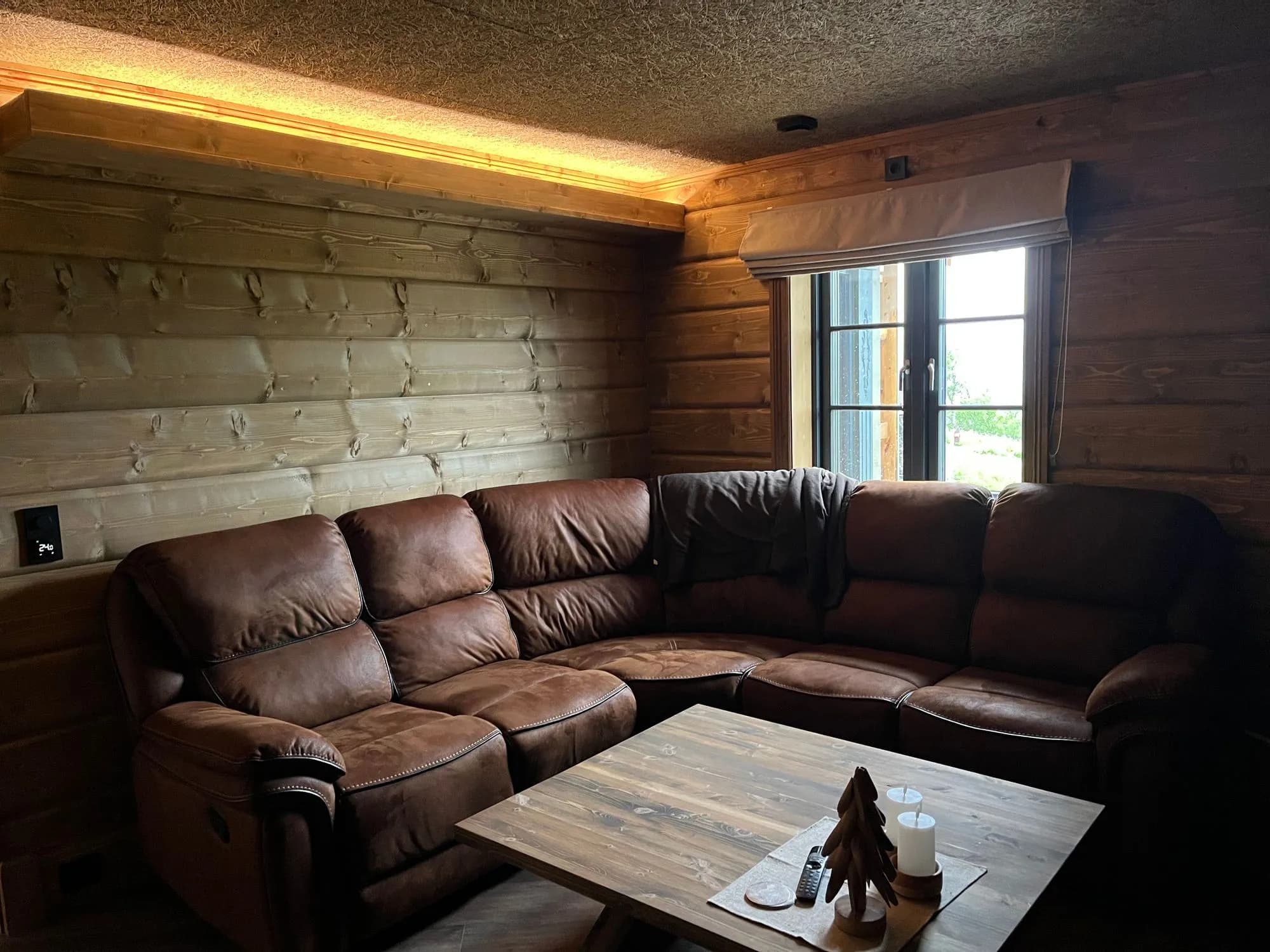 Koselig TV-stue med brun skinn-hjørnesofa, rustikt trebord og tømmervegger med indirekte lys.
