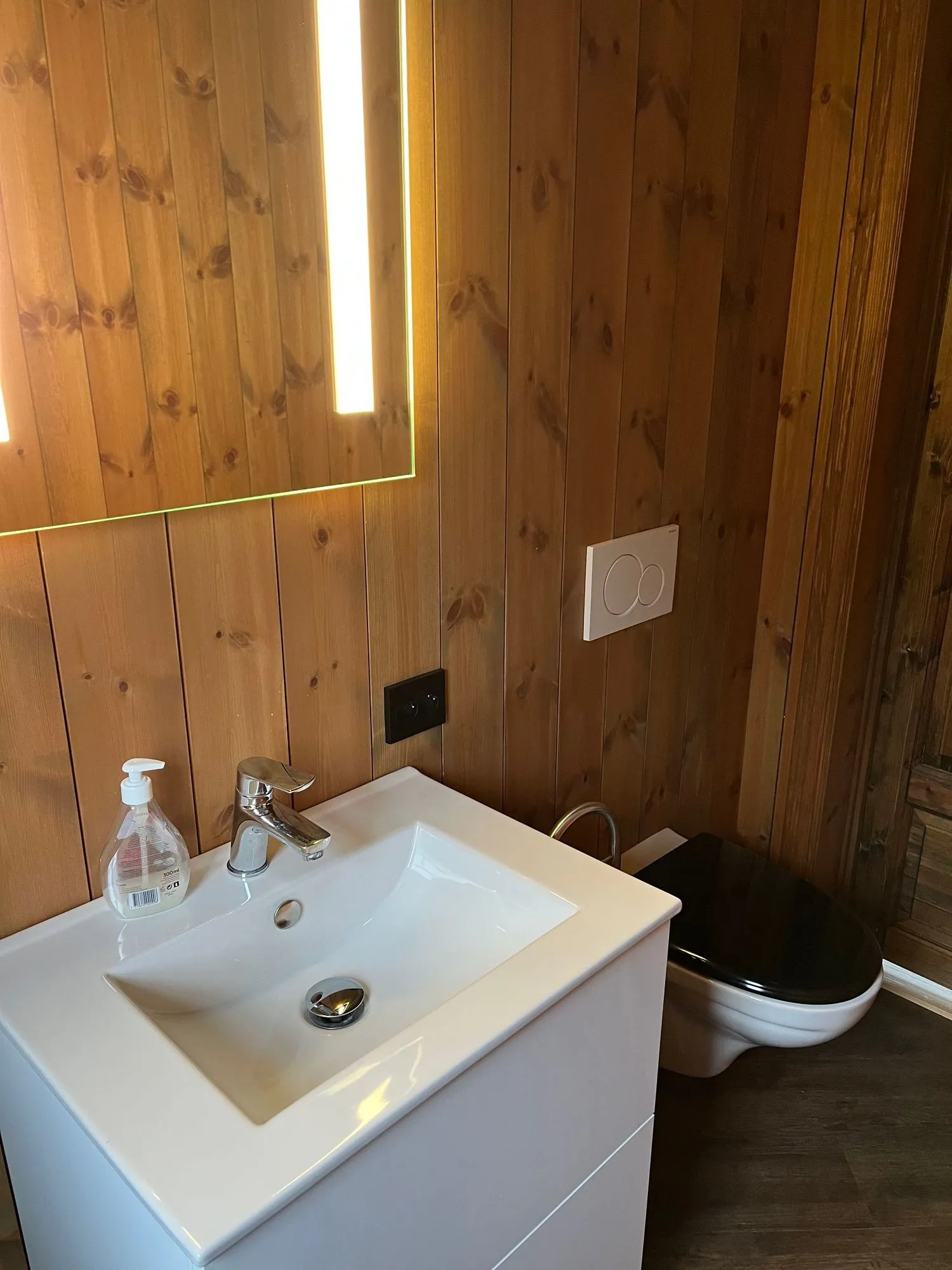 Mindre toalett med vask — furupanel-vegger, hvit servant med underskap, LED-opplyst speil og et vegghengt toalett med sort sete.